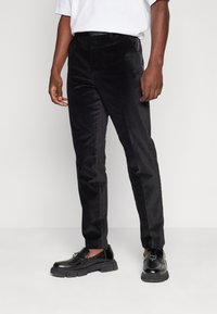 Muž v slim-fit čiernych zamatových nohaviciach a čiernych kožených loafers, stojaci na jednoliatom bielom pozadí.