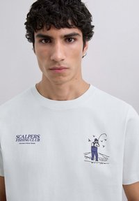 Tânăr cu părul creț și închis la culoare, purtând un tricou alb cu textul "Scalpers Fishing Club" și o grafică cu o persoană pescuind în apă.