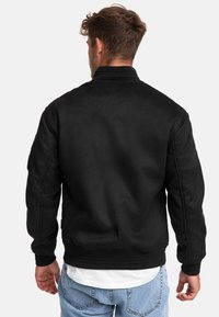 Chaqueta bomber negra con una textura suave, puños y dobladillo acanalados, detalles de costura mínimos, combinada con jeans de mezclilla azul claro y una camiseta blanca.