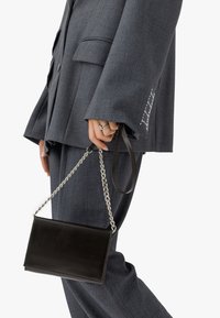 Svart läderhandväska med kedjeaxelrem, slät struktur, rektangulär form, hålls av en person i en grå oversized kostym med fickdetalj.