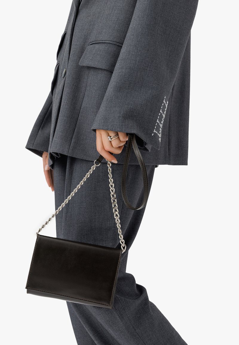 Svart läderhandväska med kedjeaxelrem, slät struktur, rektangulär form, hålls av en person i en grå oversized kostym med fickdetalj.