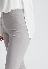 Graue eng anliegende Hose, glatter Stoff, mittelhoher Bund, flache Vorderseite, Details an den Seitennähten, kombiniert mit einer fließenden weißen Bluse.