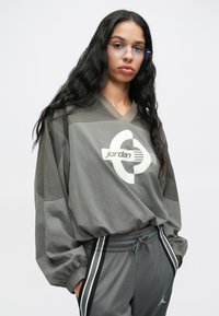 Νεαρός με μακριά σκούρα μαλλιά, φοράει διαφανή μπλε γυαλιά, έναν χαλαρό γκρι φούτερ Jordan και γκρι παντελόνια φόρμας με μαύρες και άσπρες ρίγες.