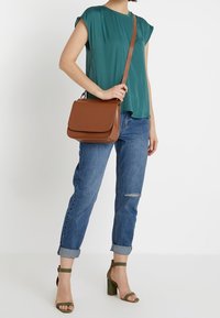Tyrkysový top s krátkým rukávem, modré džíny s ohrnutými manžetami, hnědá kožená crossbody taška, zelené sandály na blokovém podpatku s páskem kolem kotníku, mírné trhlinky na džínech.