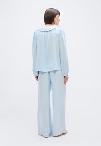 Γυναίκα όρθια ξυπόλητη, με την πλάτη προς τα εμπρός, φοράει φαρδιά ανοιχτό μπλε μπλούζα μακρυμάνικη και παντελόνι με φαρδύ πόδι, σε λευκό φόντο.