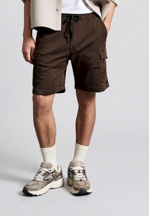 Man wearing brown cargo shorts with drawstring waist, beige sneakers, white socks, and a beige shirt with one hand in pocket.