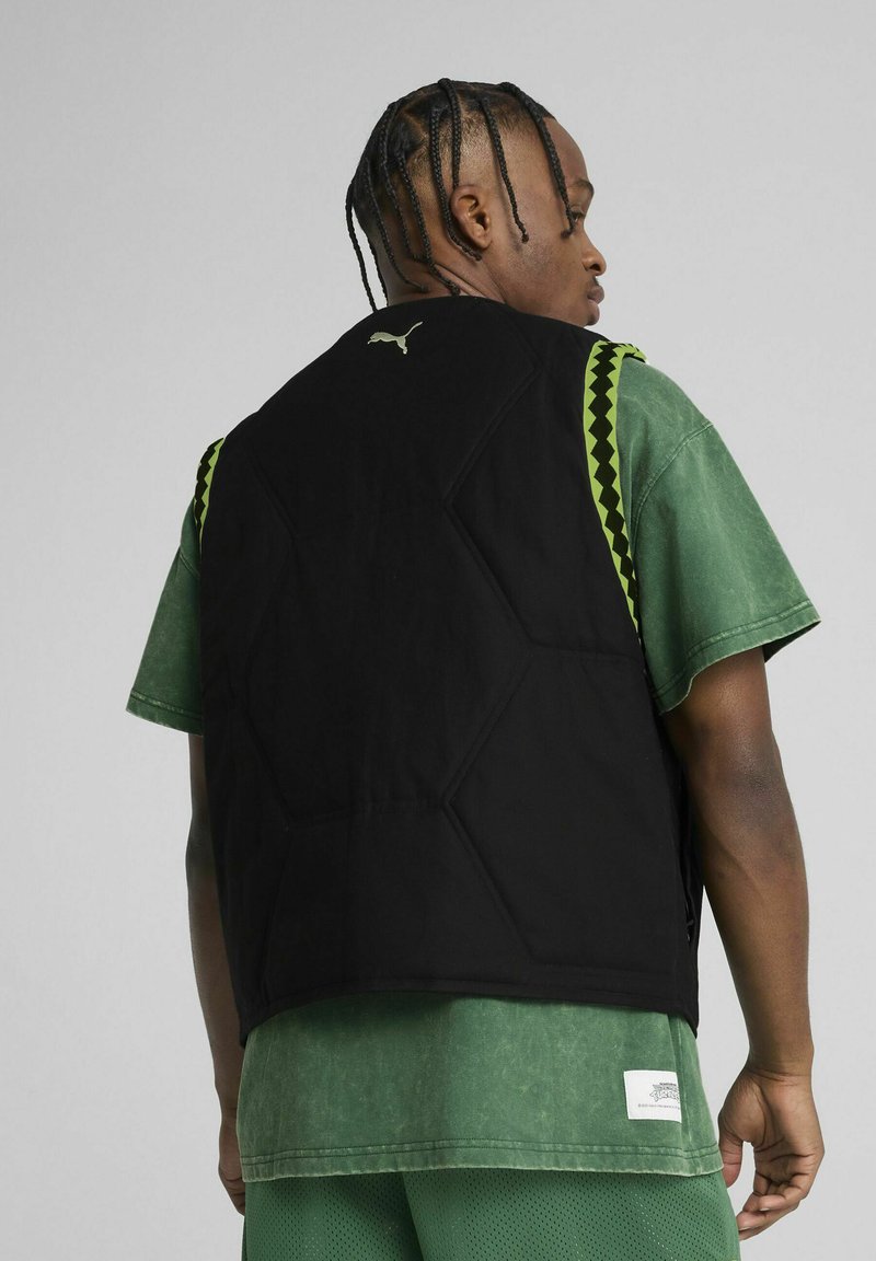Gilet rembourré noir avec un design matelassé hexagonal, accents verts sur les épaules, porté sur un t-shirt vert décoloré. Logo présent au niveau du col.