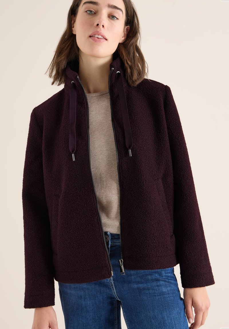 Donna con capelli marroni fino alle spalle, che indossa un giubbotto in pile viola scuro sopra un maglione beige e jeans blu, in piedi su uno sfondo neutro.