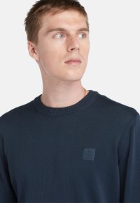 Marineblauwe gebreide trui met een geribde halslijn, voorzien van een klein vierkant logo-patch aan de linkerkant en een gladde textuur.