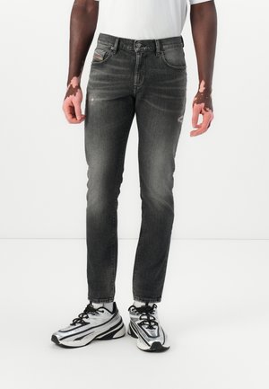 Homme portant un jean slim noir délavé et des baskets argentées et noires, debout devant un fond blanc.
