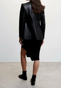 Femme aux longs cheveux portant une veste en cuir noire, une jupe noire asymétrique, des chaussettes noires et des chaussures noires, debout sur un sol gris contre un mur blanc.