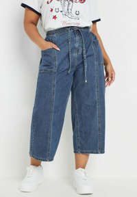 Denim culotte pants in medium blue with a drawstring waist, two side pockets, and contrasting stitching. Worn with white sneakers.
