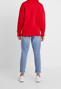 Röd sweatshirt med avslappnad passform och lång ärm, i kombination med ljusblå denimjeans och vita sportskor.