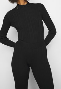 Bodysuit noir à côtes avec manches longues et col montant, présentant un design ajusté et une texture douce. Taille sans couture pour un confort optimal.
