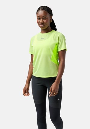 Neon žalia sportinė viršūnė su trumpomis rankovėmis, tekstūruota medžiaga ir šoniniu tinkliniu paneliu, derinama su juodais leggingais su akcento detalėmis.