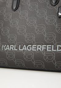 Juodas tekstūruotas odinis krepšys su pasikartojančiu baltu akinių kontūro dizainu ir ryškiu pilku "KARL LAGERFELD" logotipu apačioje.