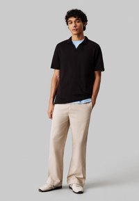 Polo shirt nero a maniche corte con scollatura a V, abbinato a pantaloni a gamba larga beige chiaro. Modello che indossa sneaker bianche. Tessuto dalla texture liscia.