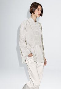 Jasnobeżowa, teksturowana bluzka z długim rękawem, z wysokim kołnierzem i pionowymi paskami, w zestawieniu z kremowymi, luźnymi spodniami.