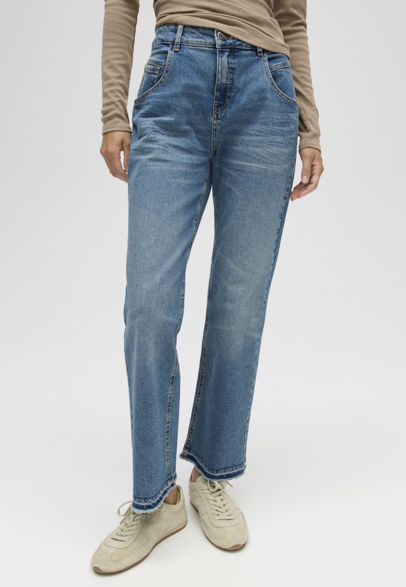 Persoon draagt lichtblauwe rechte jeans met rafelige zoom, beige top met lange mouwen en gebroken witte sneakers tegen een effen achtergrond.