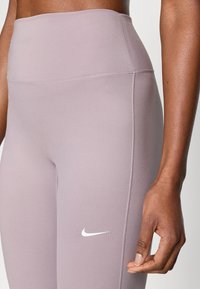 Leggings de sport violet clair avec une taille haute, une texture lisse, et un logo Nike blanc sur le côté gauche. Caractérisées par des coutures plates.