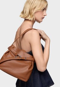 Brązowa skórzana torebka o miękkiej fakturze, luźnym fasonie i metalowych elementach. Pasek jest regulowany i przymocowany u góry.
