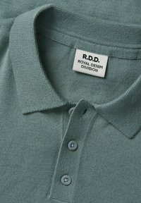 Gros plan d'un polo tricoté vert avec un col, trois boutons et une étiquette blanche portant l'inscription R.D.D. Royal Denim Division.
