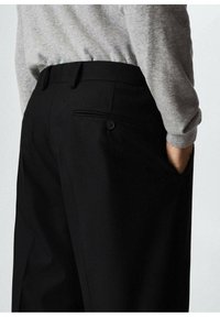 Zwarte broek met een tailored fit, met een achterzak en knoopdetail. De stof lijkt glad; de tailleband is plat en discreet.