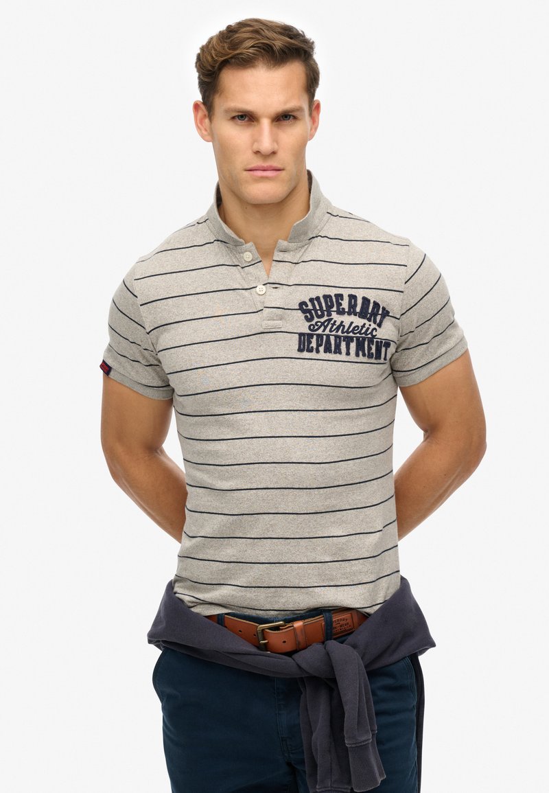 Grå polo shirt med marineblå striber, krave med to knapper og lille broderet logo på brystet. Bæres med marineblå bukser og brunt bælte.