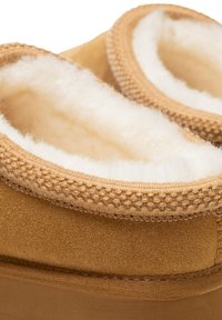 Zapatillas de ante marrón con un suave forro de vellón blanco y costuras trenzadas en las aberturas, con un exterior liso y texturizado.