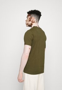 Olivgrünes T-Shirt mit kurzen Ärmeln, Rundhalsausschnitt, aus softem Baumwollstoff, hat einen geraden Schnitt, die Rückansicht zeigt ein Etikett am Ärmel.