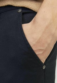 Navy pants with a textured fabric, featuring a side pocket and a visible metal button near the opening. Close-up of hand in the pocket.