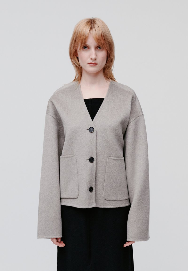 Hellgrauer Wollmischcardigan mit V-Ausschnitt, vier schwarzen Knöpfen und zwei vorderen Taschen. Lockere Passform mit weiten Ärmeln.