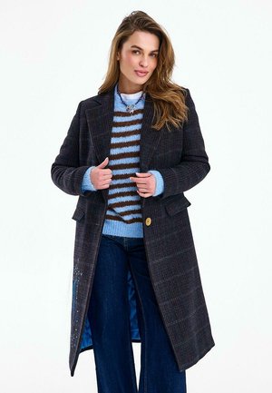 Kobieta poprawiająca ciemny kraciasty długi płaszcz na sobie niebieski i brązowy sweter w paski, nosząca szerokie spodnie dżinsowe i naszyjnik, stojąca na tle białego tła.