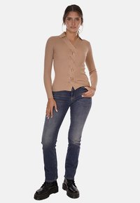 Maglietta lunga a maniche con costine color beige, scollatura a V e dettaglio anteriore attorcigliato, abbinata a jeans aderenti blu scuro e stivali neri con lacci.