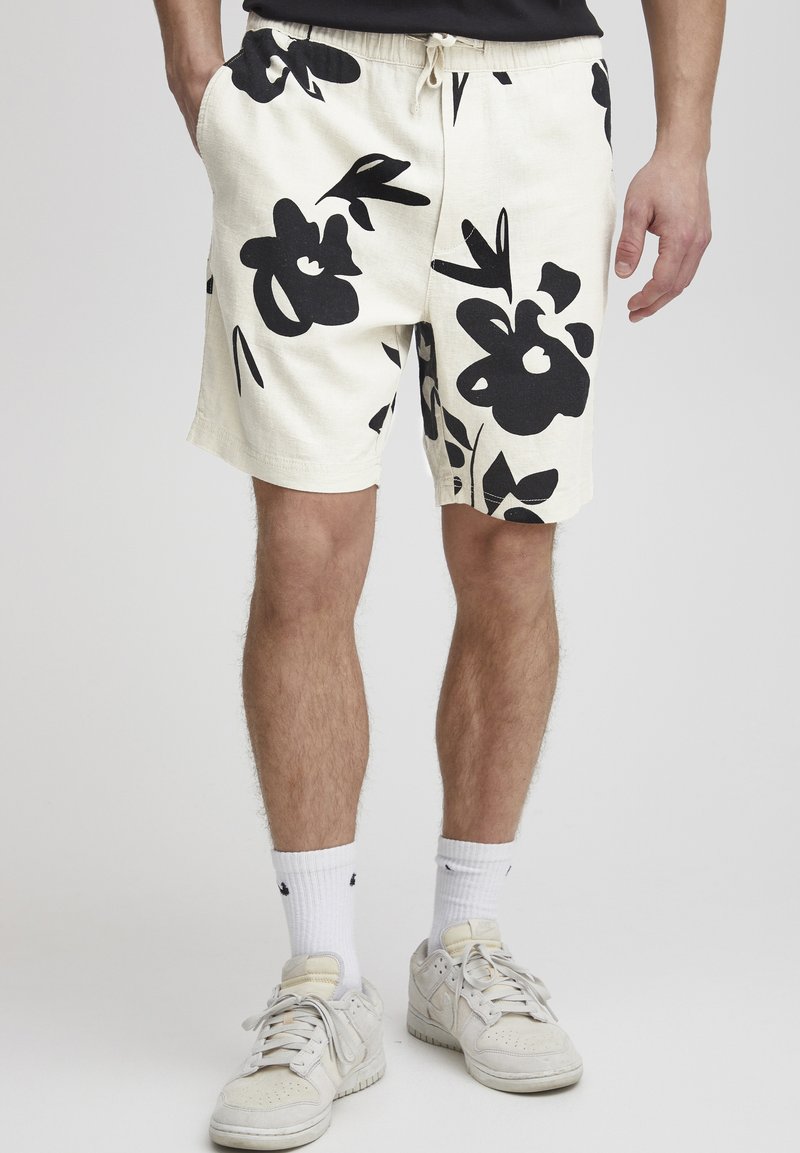 Sorte blomstrete shorts i et lyst beige stoff, med elastisk midjebånd og snor. Sammen med hvite sokker og lyse joggesko.