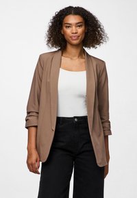 Mujer con cabello rizado lleva una chaqueta marrón claro con mangas remangadas, camiseta blanca y pantalones negros de cintura alta, de pie contra un fondo blanco.