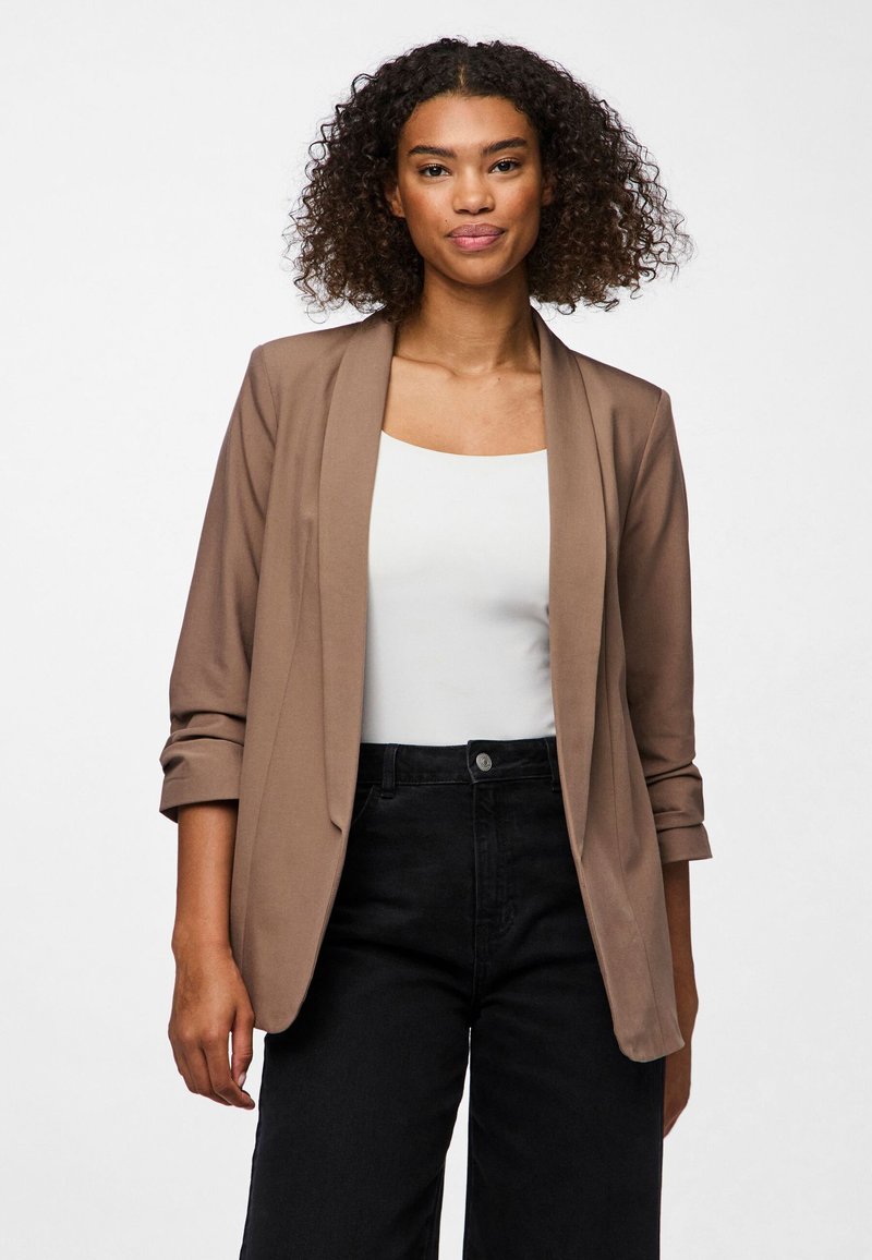 Mujer con cabello rizado lleva una chaqueta marrón claro con mangas remangadas, camiseta blanca y pantalones negros de cintura alta, de pie contra un fondo blanco.