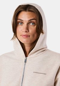 Hoodie zippé beige clair avec une texture douce, doté d'une capuche et d'un logo "SWEDEMOUNT" sur la poitrine, avec un détail de fermeture éclair argenté.