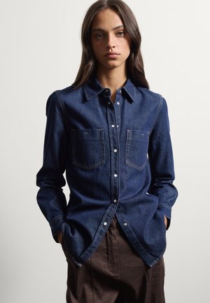Giovane donna che indossa una camicia di jeans blu scuro con bottoni e due tasche sul petto e pantaloni marroni, in piedi con le mani nelle tasche.