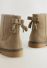 Hnědé suede kotníkové boty se sraženým podpatkem, s mašlemi a třásněmi na zadní straně. Texturovaný povrch a kontrastní detail podešve.
