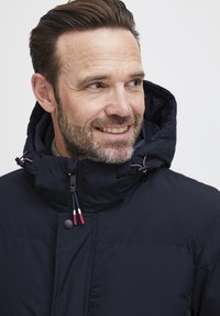 Mørkeblå pufferjakke med høj krave og justerbar hætte. Har lynlåslukning med farvede træk og en quiltet tekstur.