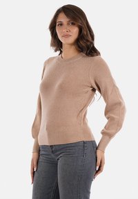 Maglione beige realizzato in tessuto a maglia texturizzata, con collo tondo, maniche lunghe e orlo aderente. Indossato con jeans di denim scuri.