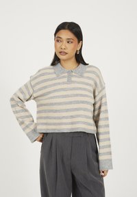 Kobieta z długimi ciemnymi włosami, ubrana w beżowy i szary sweter w paski z kołnierzykiem oraz spodnie o wysokim stanie w ciemnoszarym kolorze, stojąca z jedną ręką na biodrze.