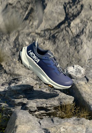 Chaussure de running pour homme avec une tige en synthétique bleue, une semelle intermédiaire blanche et une semelle extérieure noire. Design texturé avec des lacets réfléchissants et des détails de logo.