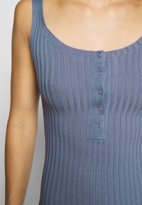 Ribbad, utan ärmar topp i mjukt blått tyg med rundad halsringning och fem knappar nedanför mittfronten.