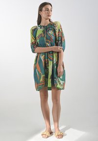 Modelka stoi pewnie w kolorowej sukience w wzór, z balonowymi rękawami, zestawiona z teksturowanymi sandałami, na jasnym tle.
