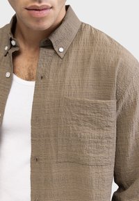 Brown textured button-up shirt with a collar, single chest pocket, and visible stitching details. Worn over a white undershirt.