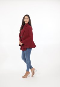 Capa de burgundy con una silueta estructurada, cuello alto y detalle de cinturón, combinada con jeans ajustados azules y sandalias de tacón transparente.