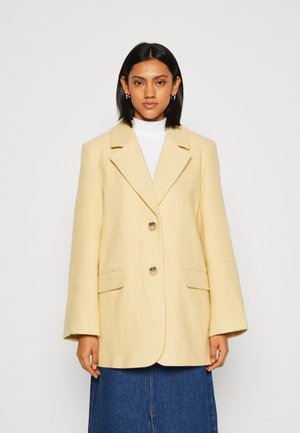 Κίτρινο σακάκι με υφή και γιακά με γωνίες, κλείνει με δύο κουμπιά και έχει μπροστινές τσέπες, φορεμένο πάνω από λευκή μπλούζα με λαιμόκοψη και τζιν φούστα.