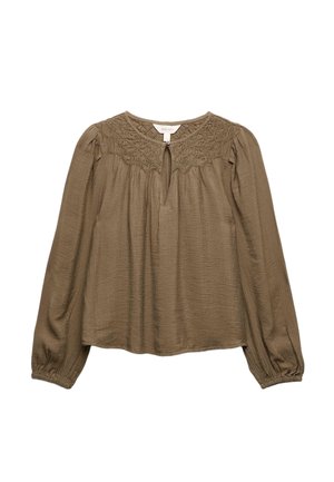 Kvinde med krøllet mørkt hår, iført brun bluse, lagdelte sølvsmykker, store øreringe, blå jeans og en ring, siddende og kiggende til siden.